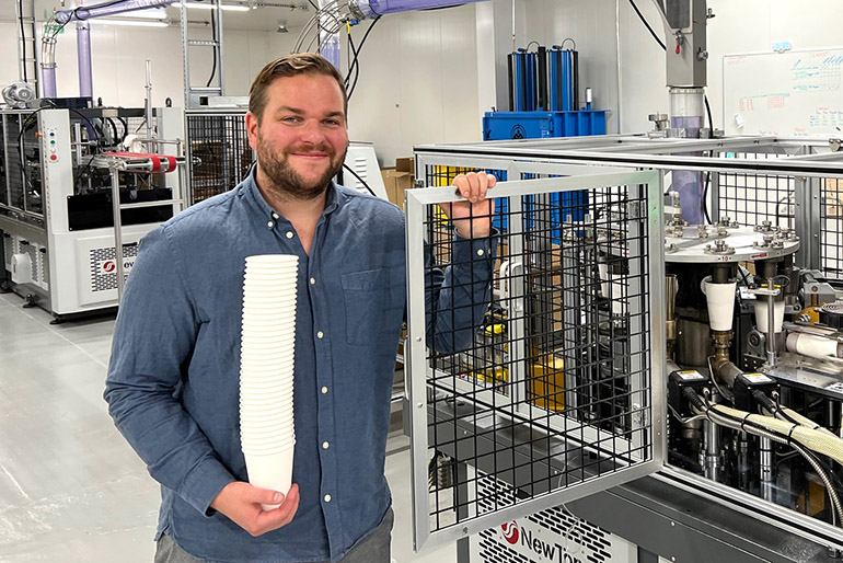 Miika Hämäläinen hymyilee ja katsoo kohti kameraa kartonkikuppeja valmistavan koneen vierellä.