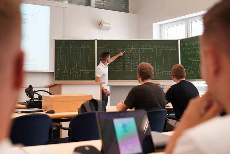 Oppilaat kuuntelevat pulpettien äärellä, kun opettaja opettaa ja kirjoittaa samalla liitutaululle.