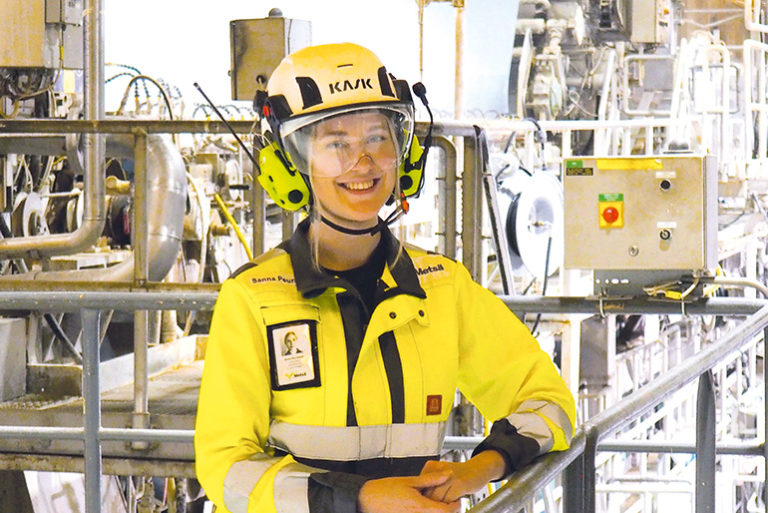 Sanna Peuralampi keltaisessa työhaalarissa, kypärä ja kuulosuojaimet päässään ja katsoo hymyillen kohti kameraa.