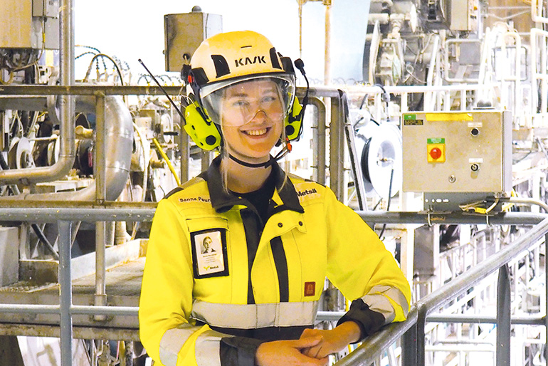 Sanna Peuralampi keltaisessa työhaalarissa, kypärä ja kuulosuojaimet päässään ja katsoo hymyillen kohti kameraa.