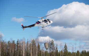 Helikopteri lentää metsän yllä ja levittää lannoitetta vaijerin päässä roikkuvasta säiliöstä.