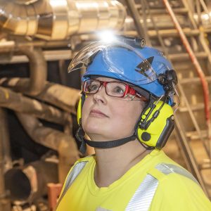 Sini Kurki katsoo tehtaalla huollettavaa laitetta suojavarusteet kuten kypärä, suojalasit ja kuulosuojaimet päässään,.